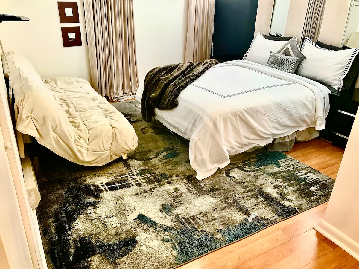 The bedroom features a spacious queen bed covered with white linens and accent pillows. A cozy futon is positioned nearby on a patterned area rug, adding extra sleeping space. Natural light filters through sheer curtains, creating a warm and inviting atmosphere.