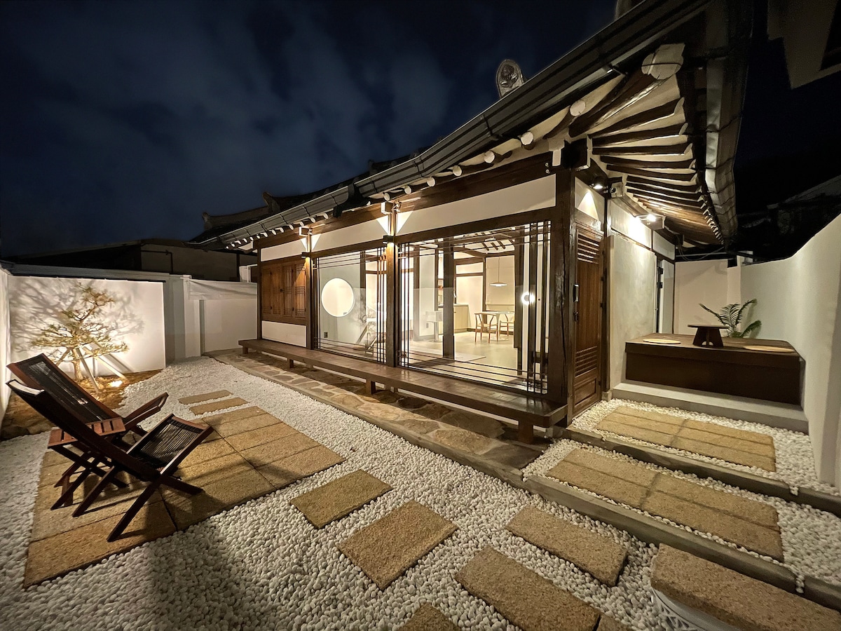 The traditional Hanok structure is illuminated at night, showcasing wooden beams and sliding glass doors. A serene outdoor space features gravel pathways and a wooden deck, accompanied by lounge chairs. Soft lighting highlights the surrounding plants and contributes to a peaceful ambiance.