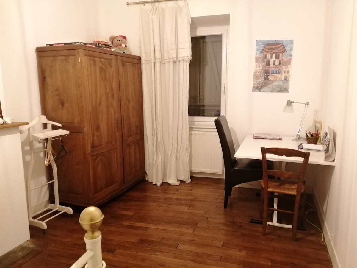 A tidy room features a wooden wardrobe and a window dressed with white curtains. A desk and chair are positioned against one wall, accompanied by a small bookshelf. A wall art piece adds a touch of color to the neutral decor.