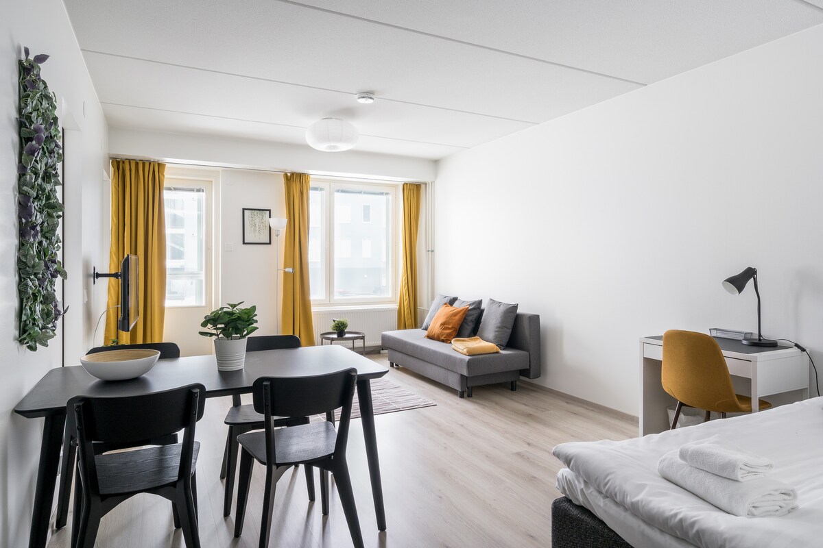 A bright living area features large windows with yellow curtains, allowing natural light to fill the space. A dining table is accompanied by black chairs, while a gray sofa offers additional seating. A desk with a lamp is positioned beside the bed, creating a functional workspace.