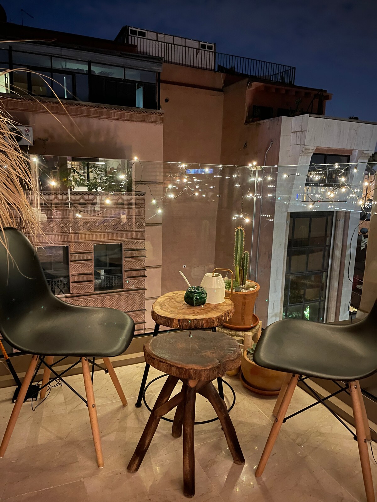A cozy balcony setting features two modern chairs beside a wooden stool adorned with a small plant and refreshment. Soft, twinkling fairy lights illuminate the area, enhancing the view of the surrounding buildings against the night sky.