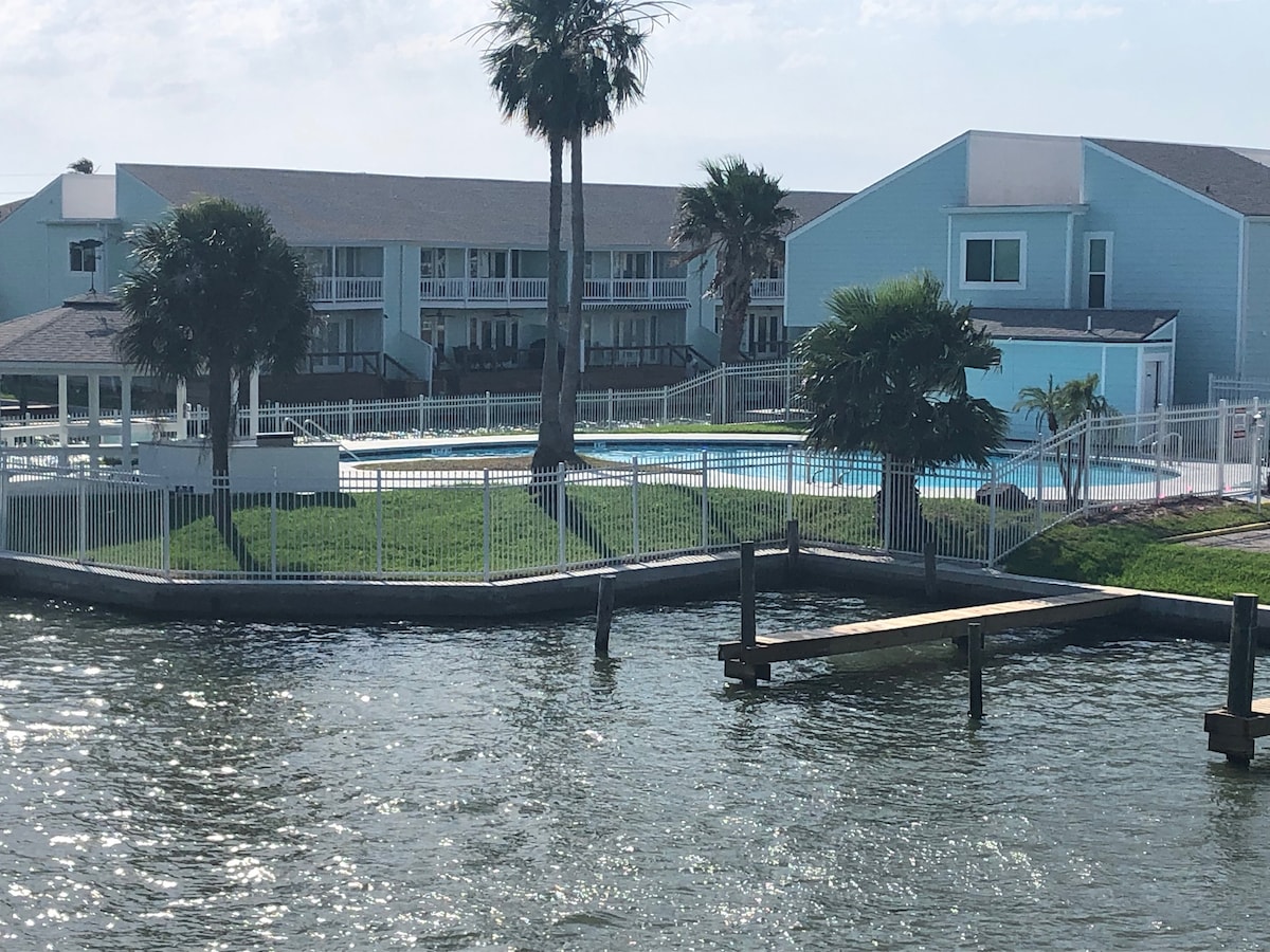 A view of the swimming pool surrounded by green grass and palm trees, located within a fenced area. The pool area is complemented by a nearby residential building with light blue siding, set against a bright sky. Water ripples reflect the sunlight.