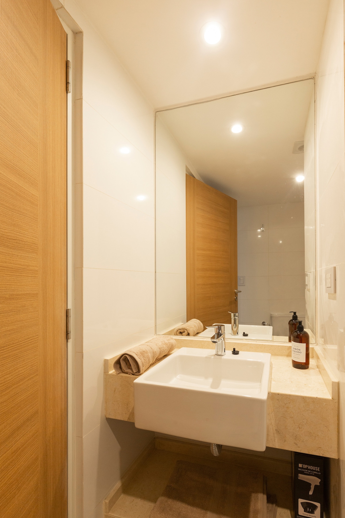 A modern bathroom features clean lines and neutral tones. A sleek sink is positioned on a light-colored countertop with a rolled towel beside it. Large mirrors reflect the bright lighting, and a wooden door is visible in the background.