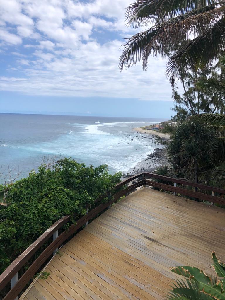 A wooden deck extends out towards the ocean, framed by lush greenery and palm trees. The sea is visible with gentle waves lapping against the shore, while the coastline stretches into the distance under a partly cloudy sky.