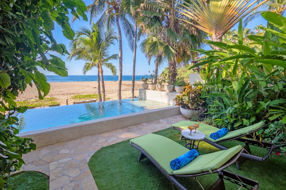 A serene outdoor area features two green lounge chairs beside an infinity pool with ocean views. Lush tropical plants surround the space, enhancing the connection to nature. The sandy beach is visible in the distance, creating a peaceful seaside ambiance.