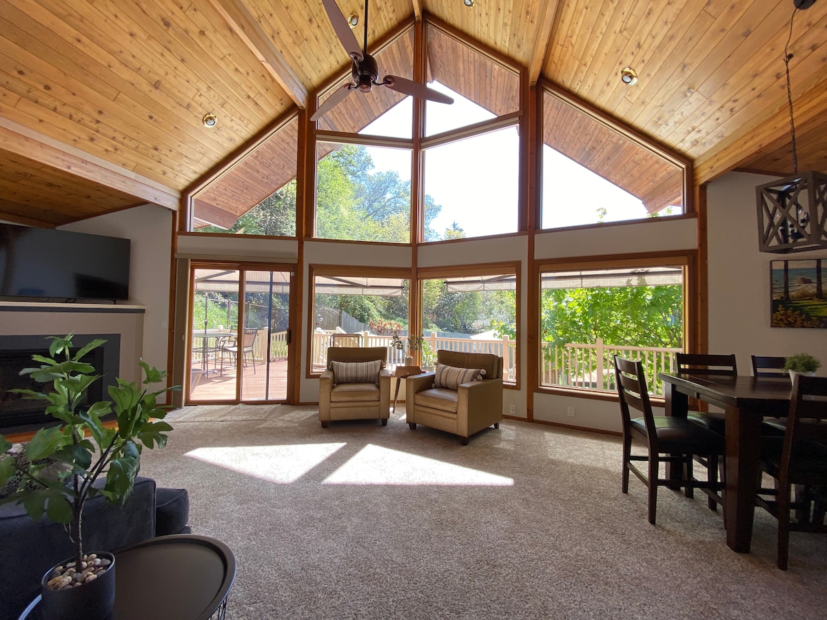 The open-concept great room features a wall of glass that brings in natural light, framed by wooden cathedral ceilings. Two plush armchairs are positioned for relaxed seating, with a dining area visible in the background. The space connects seamlessly to the outdoor deck.