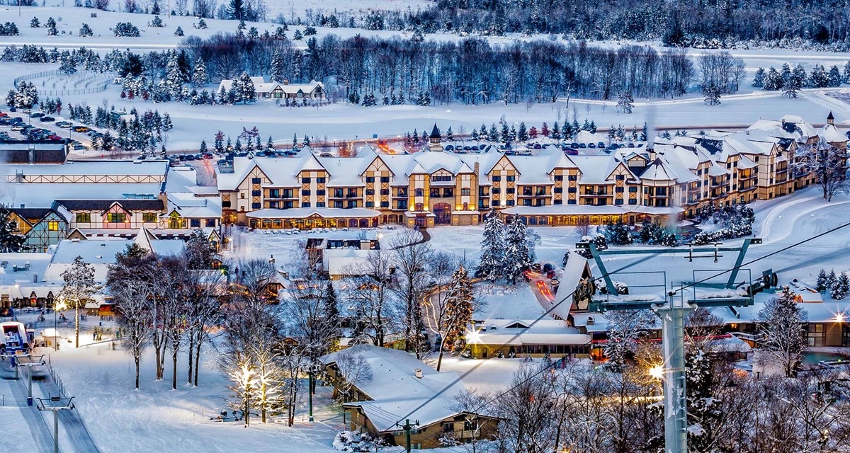 A winter landscape features a charming cluster of buildings blanketed in snow, surrounded by a tranquil forest. Soft lights illuminate the pathways, while a ski lift is visible in the foreground. The serene atmosphere highlights the beauty of the snowy setting.