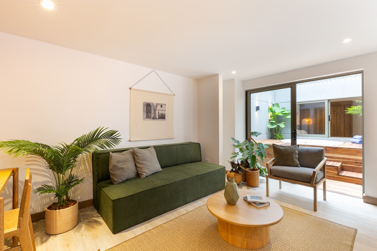 A modern living area features a green sofa with cushions, paired with a wooden side chair. A round coffee table sits in the center, while light pours in through large glass doors that open to a small outdoor space with plants.