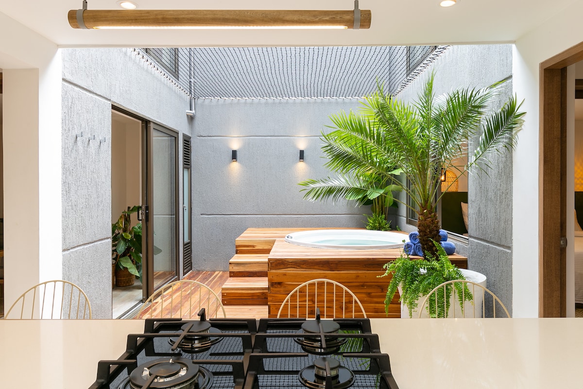 An open kitchen is highlighted by sleek appliances and a view of a private jacuzzi surrounded by lush greenery. Natural light floods the space through expansive sliding doors, creating a seamless connection to the courtyard's wooden deck.