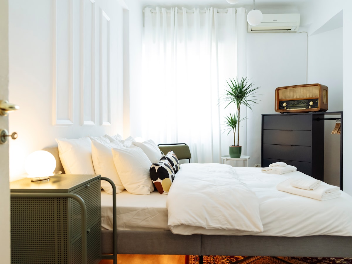A well-lit sleeping area features a spacious bed adorned with multiple pillows and soft linens. A decorative plant adds a touch of greenery, while a vintage radio sits on a dark dresser. Light curtains frame a window, allowing natural light to fill the room.