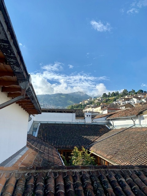 A panoramic view captures the terracotta rooftops of Quito's old town, framed by verdant hills and a clear blue sky. The image illustrates a blend of traditional architecture, with the mountainous landscape providing a tranquil backdrop to the urban scene.