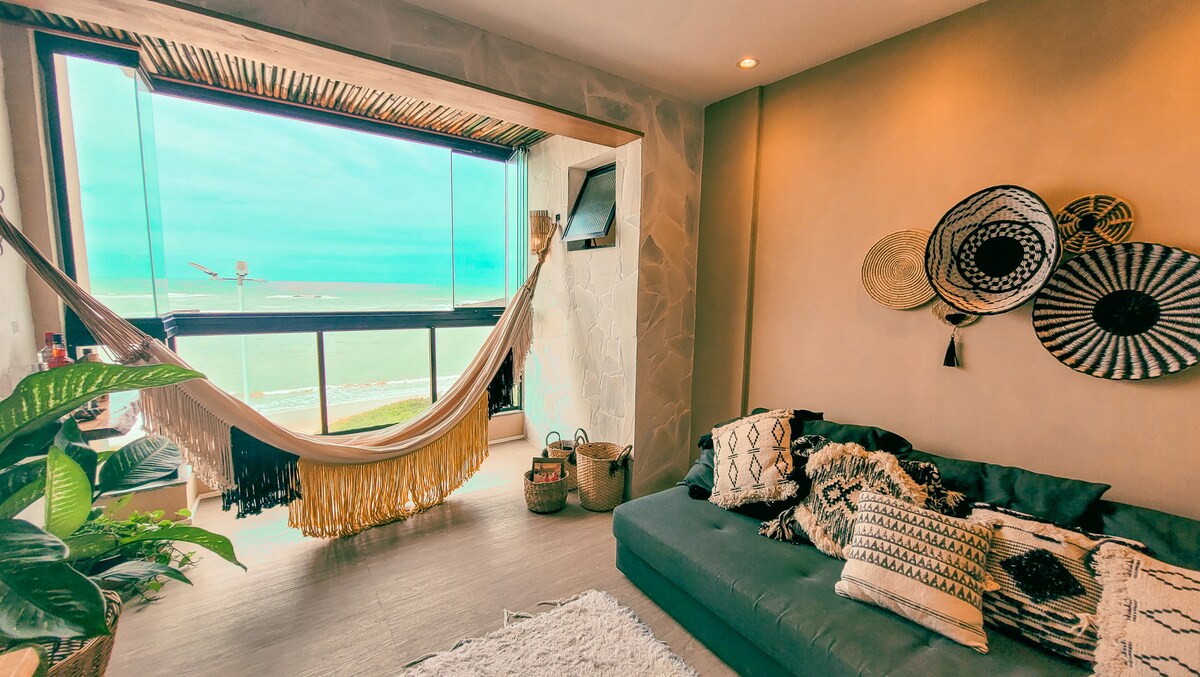A bright and airy living space is enhanced by large windows providing a view of the sea. A cozy gray couch is adorned with various cushions, while a hammock is suspended nearby. Decorative wall accents add texture, and greenery from nearby plants brings life to the area.