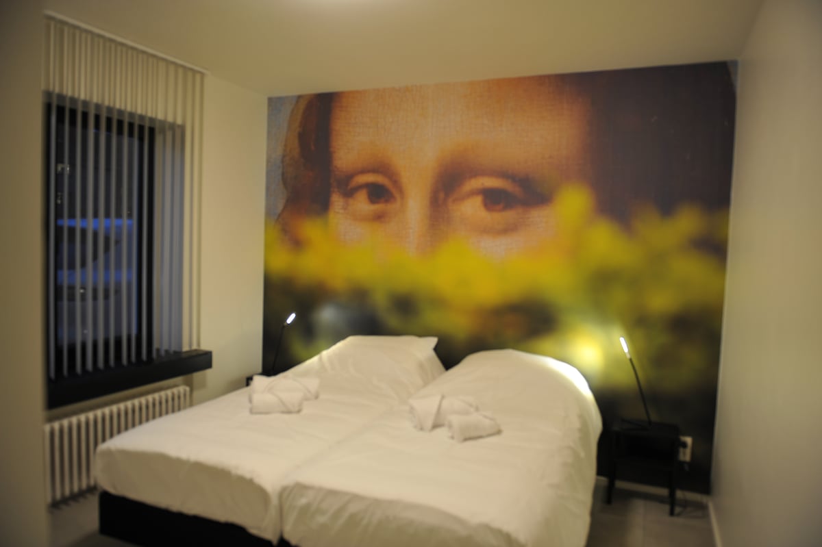 The bedroom features two spacious boxspring beds arranged side by side. Soft bedding is neatly arranged, complemented by bedside lamps. A large window with vertical blinds allows natural light to enter the room, casting a warm glow on a colorful accent wall.