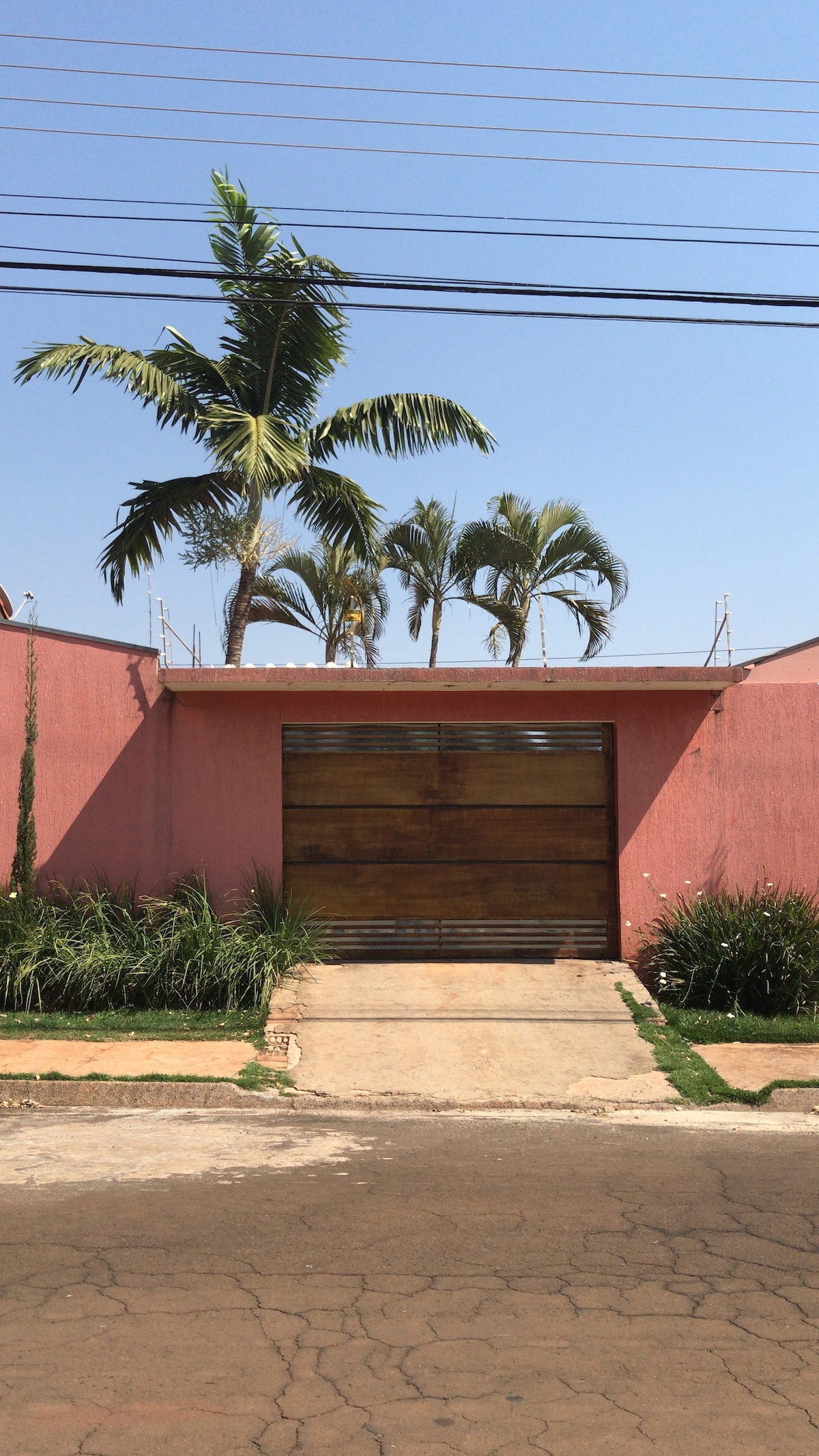 A pink exterior wall is visible, accented by lush green grass along the pathway. A large wooden gate, partially closed, leads to a private entrance. Tropical palm trees stand tall against a clear blue sky, creating a serene atmosphere in the neighborhood.