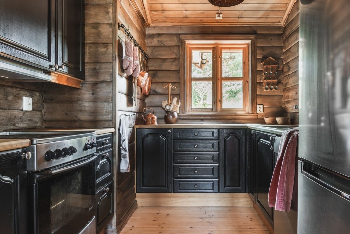 The kitchen features dark wooden cabinetry with a charming rustic feel. A window allows natural light to brighten the space, while cooking utensils and bowls are neatly arranged on the walls and countertops. Modern appliances, including an oven and refrigerator, complement the traditional design.
