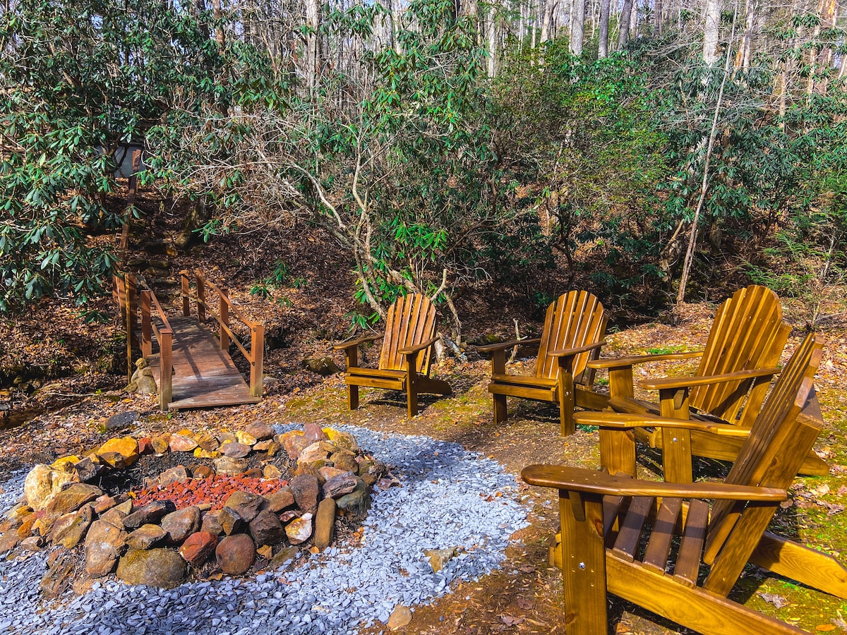 An outdoor fire pit area is surrounded by four wooden Adirondack chairs, nestled among lush greenery. The fire pit, constructed from stones, is centrally located, providing a cozy gathering spot. A wooden bridge in the background leads over the creek, enhancing the natural setting.