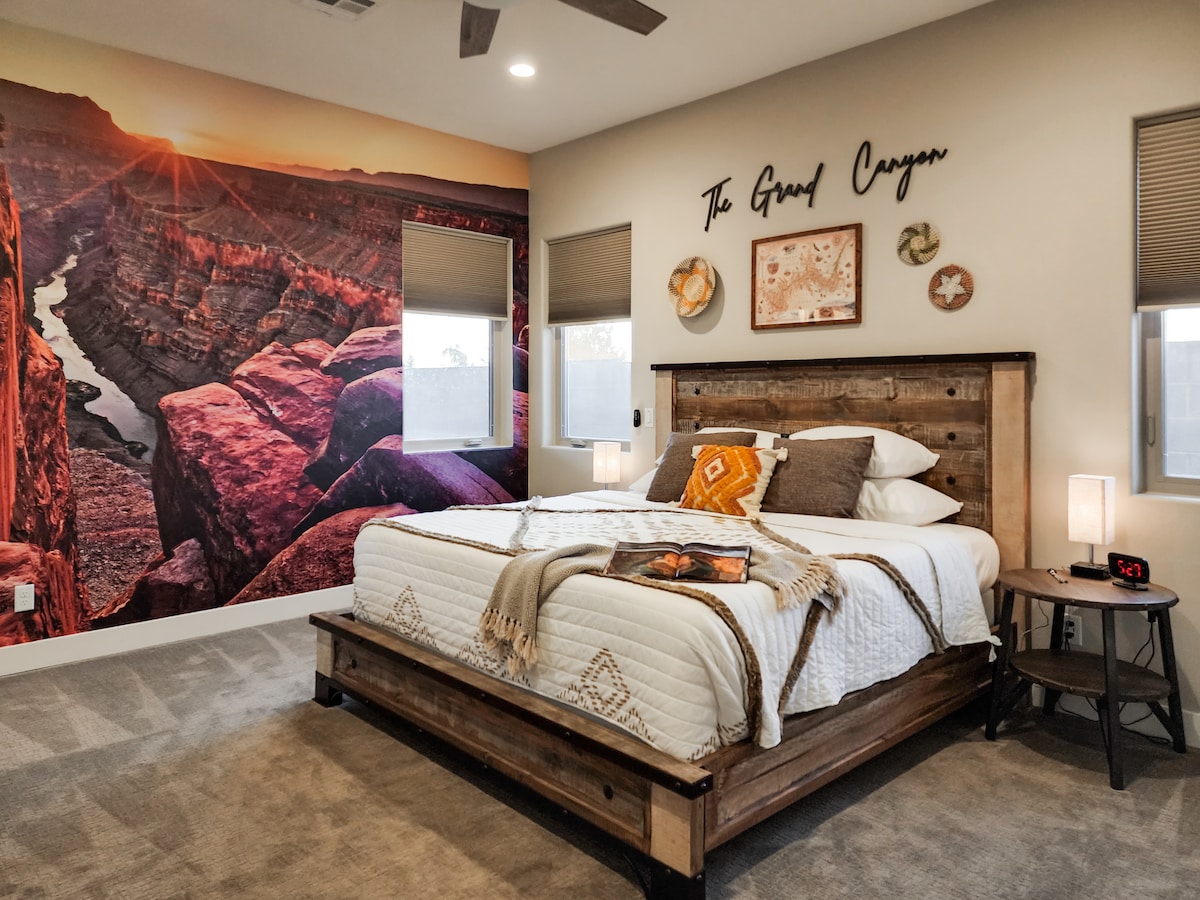 A spacious bedroom features a king bed with a quilted coverlet placed against a striking wall mural of the Grand Canyon. Soft light filters through two windows with blinds, and decorative wall art complements the rustic wooden furnishings.