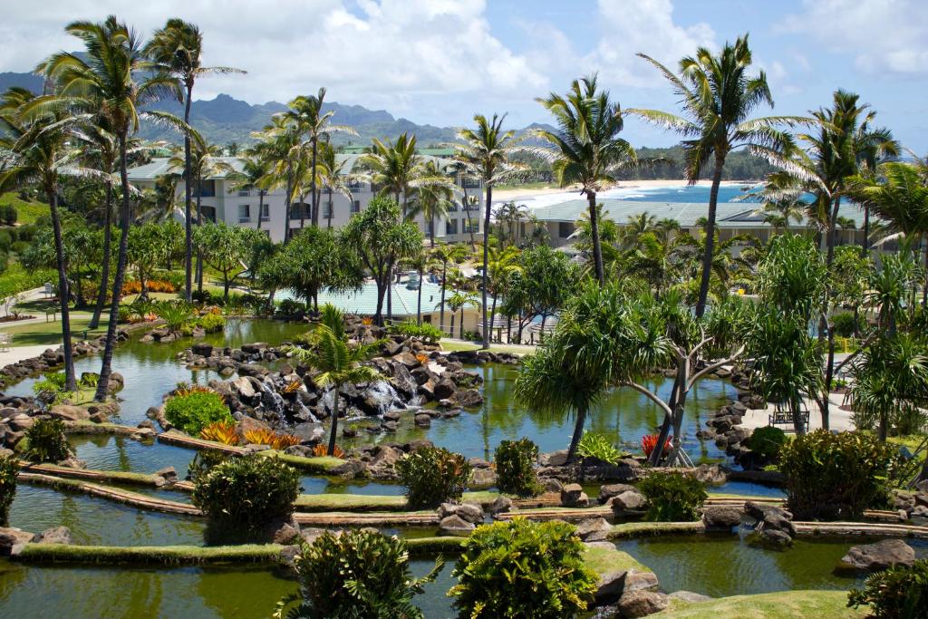 A lush garden area is depicted, featuring a tranquil pond surrounded by tropical plants and palm trees. A pool area can be seen in the background, with the resort buildings and distant mountains framing the scenery under a partly cloudy sky.