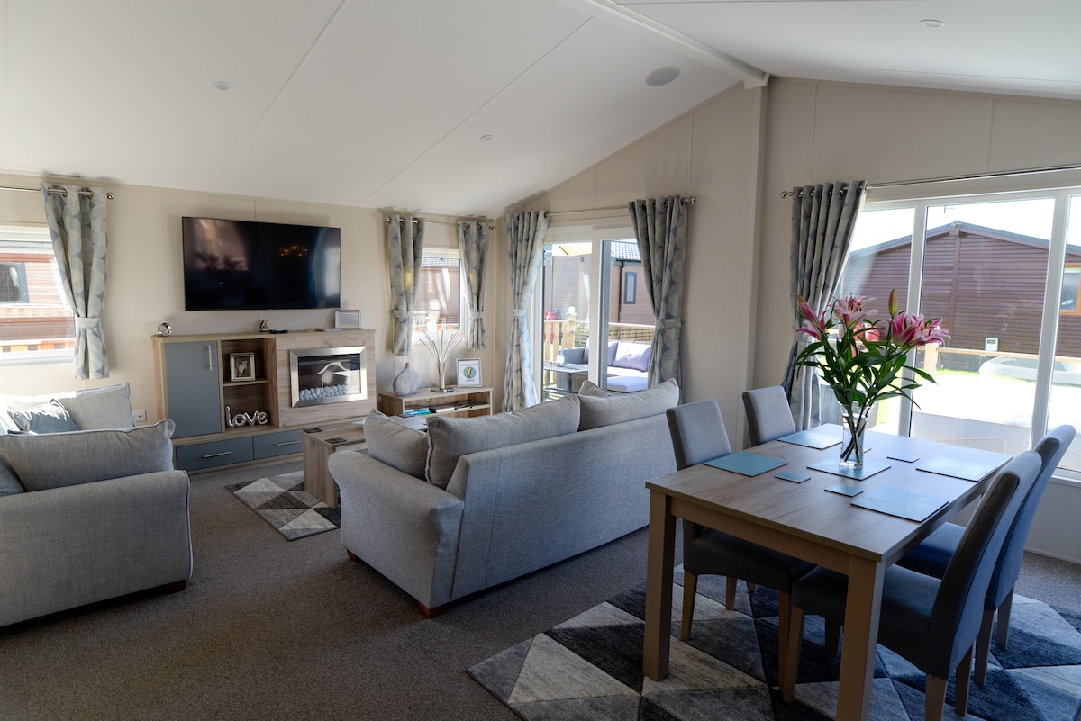 The bright and airy open-plan living area features a comfortable seating arrangement with light grey sofas and a modern television mounted above a stylish fireplace. A dining table with four chairs and a floral centerpiece enhances the inviting atmosphere, while large windows allow for ample natural light.