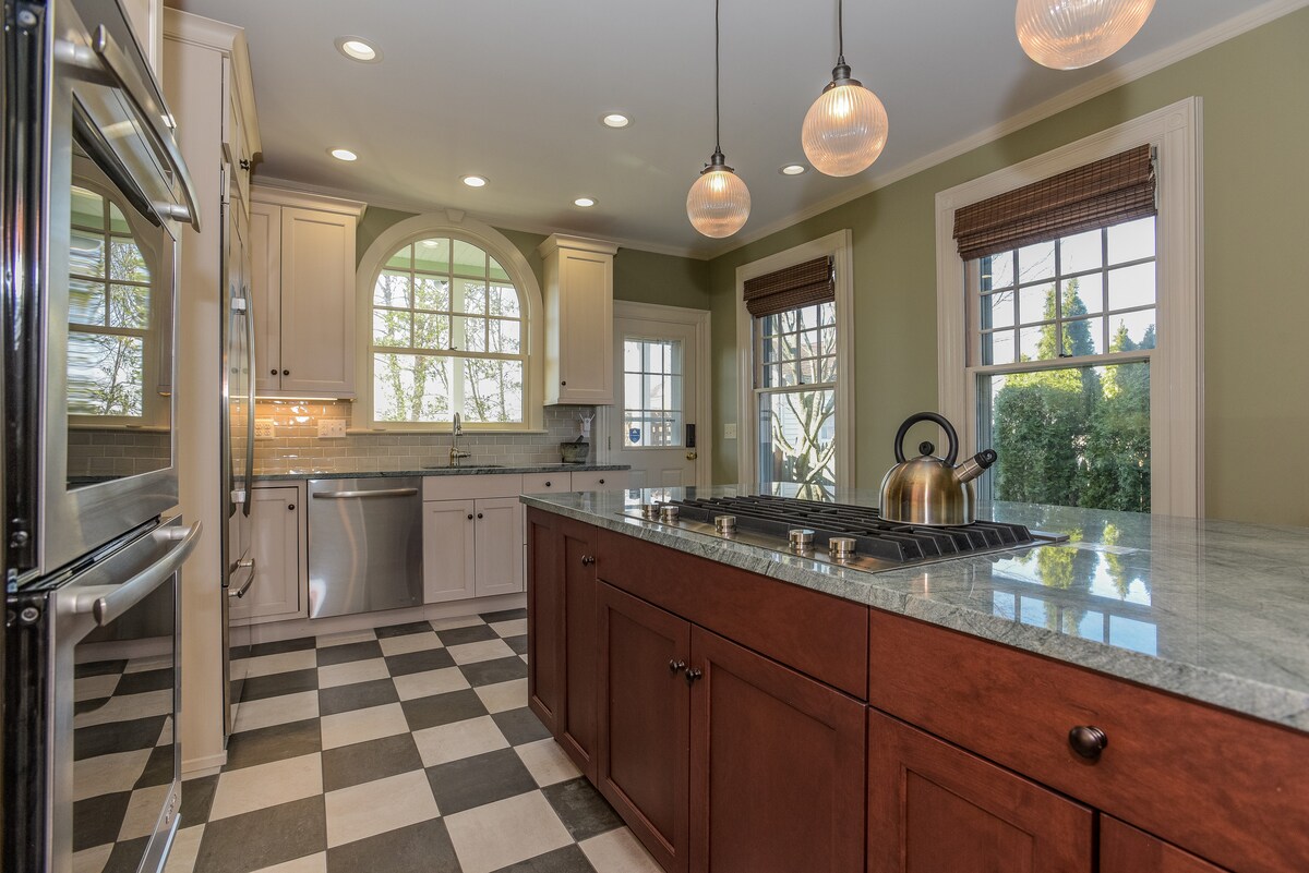 A modern kitchen is highlighted by a central island with a dark wood finish and a stovetop. Checkered flooring creates visual interest, while large windows allow natural light to fill the space, showcasing a blend of traditional and contemporary cabinetry.