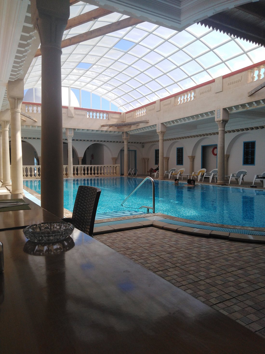 An indoor swimming pool is framed by tall columns, with sunlight filtering through a glass ceiling. Loungers are arranged around the water, creating a relaxing atmosphere. The decor is characterized by traditional elements, enhancing the serene environment.