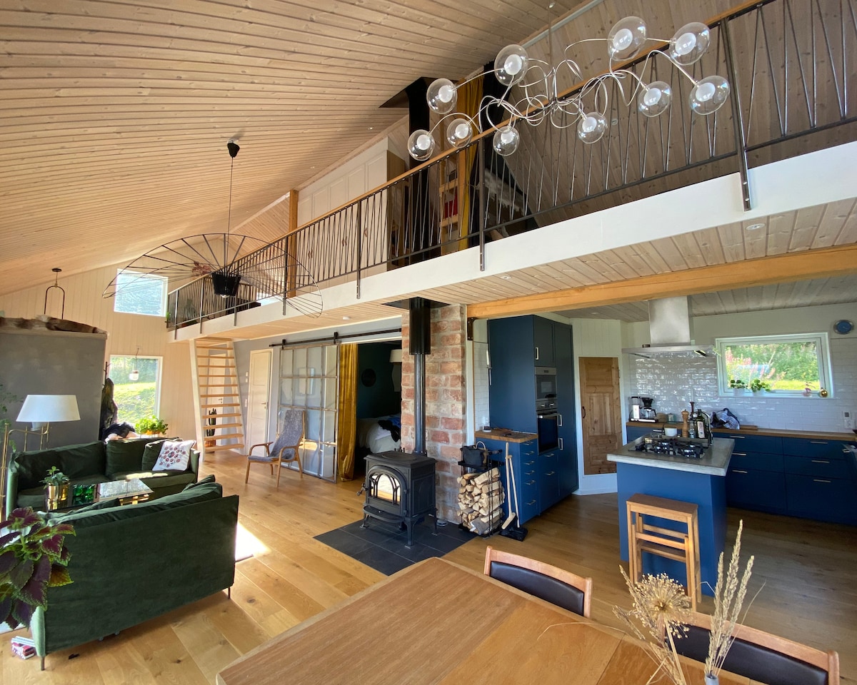 An open-plan living area features high ceilings and natural wood finishes. A cozy seating arrangement is positioned near a Jötul wood stove. A modern kitchen with a navy blue cabinetry is visible, along with a dining table that complements the warm wooden flooring.