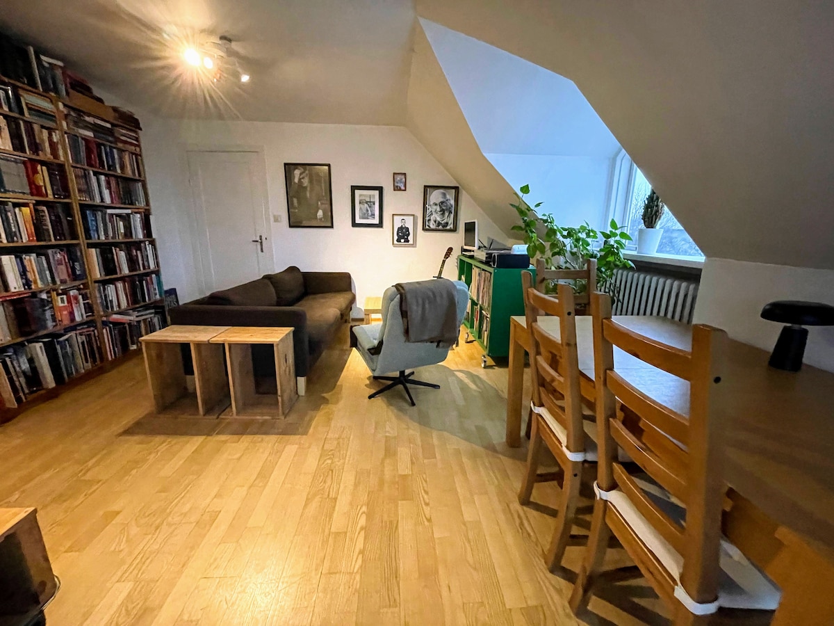 An open living area features a comfortable couch and two wooden coffee tables. A chair with a soft upholstery is present, alongside a dining table set with chairs. A well-stocked bookshelf lines one wall, with framed art and a potted plant adding interest.
