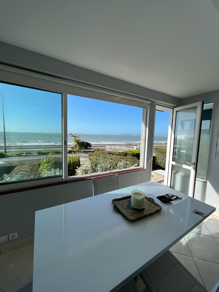 A bright dining area features large windows that offer views of the sea and the surrounding greenery. A white table is set with a candle and a small tray, allowing light and fresh air to fill the space.