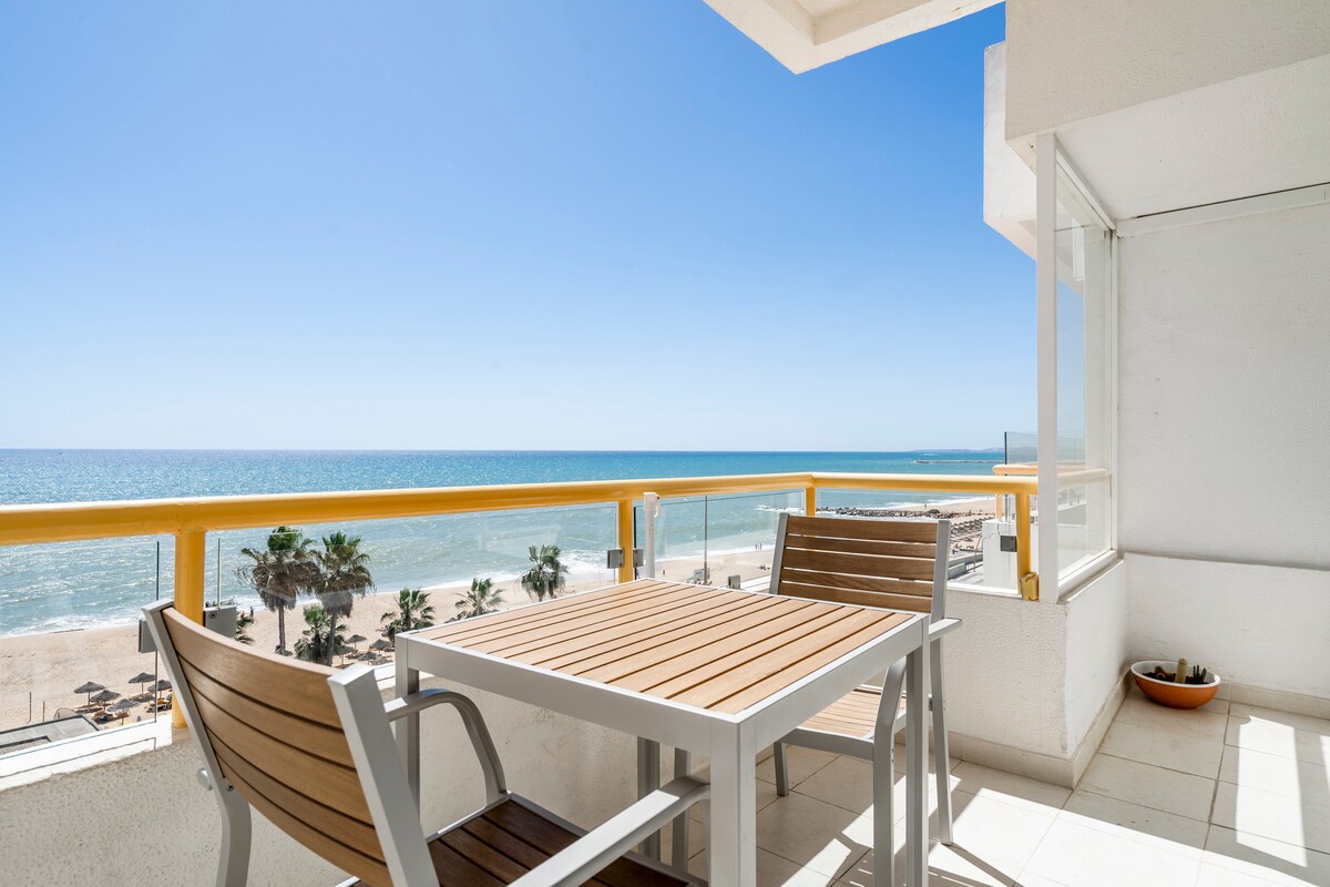 A private balcony offers an unobstructed view of the ocean and beach, with sunlight reflecting off the water. A wooden table is accompanied by two chairs, creating a space for relaxation and meals while enjoying the coastal scenery.