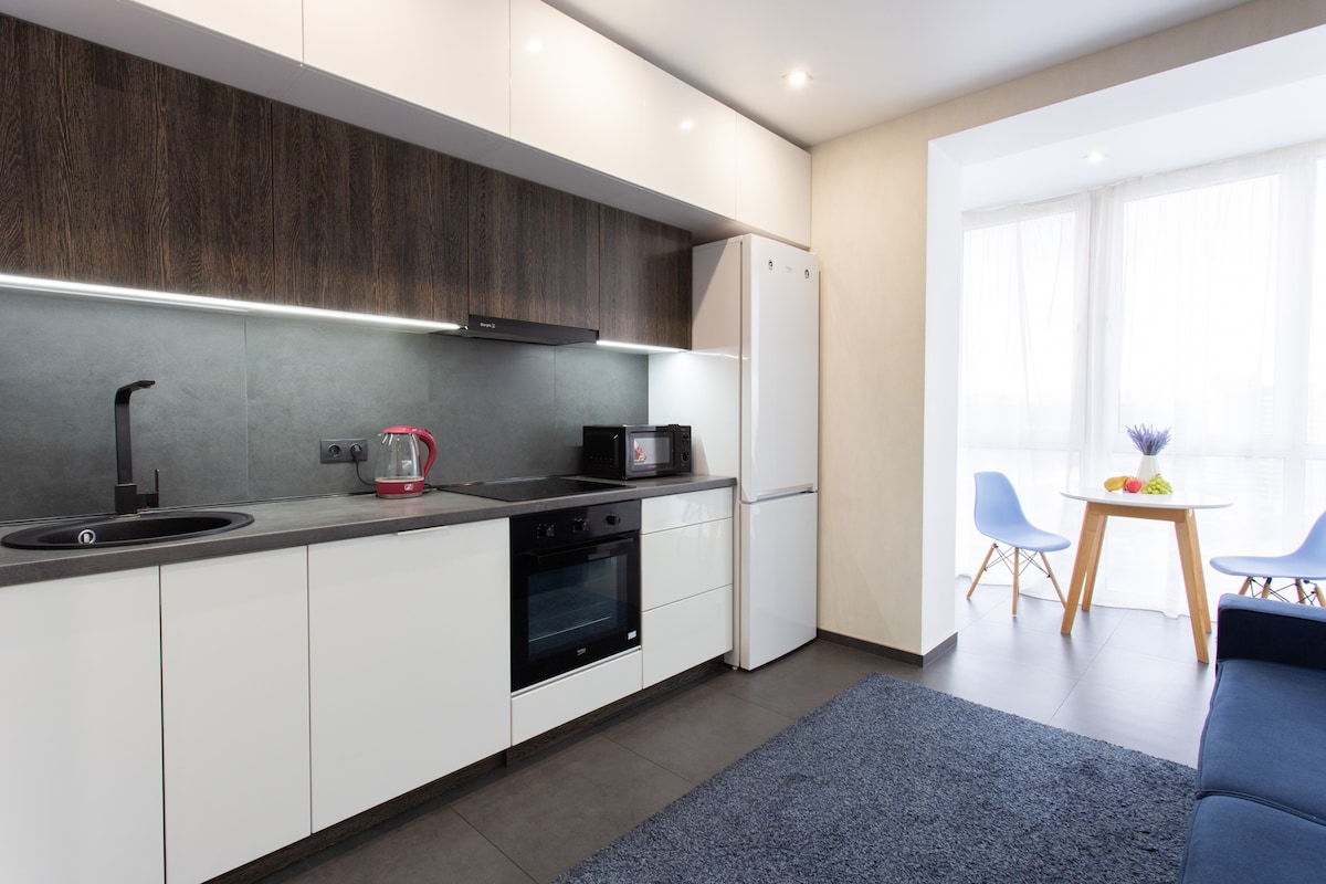 The modern kitchen area features sleek cabinetry, a built-in oven, and a stovetop, complemented by a stylish sink. Natural light filters through the window, illuminating a small dining table with two chairs. A living area with a dark sofa completes the space, creating a functional and inviting atmosphere.