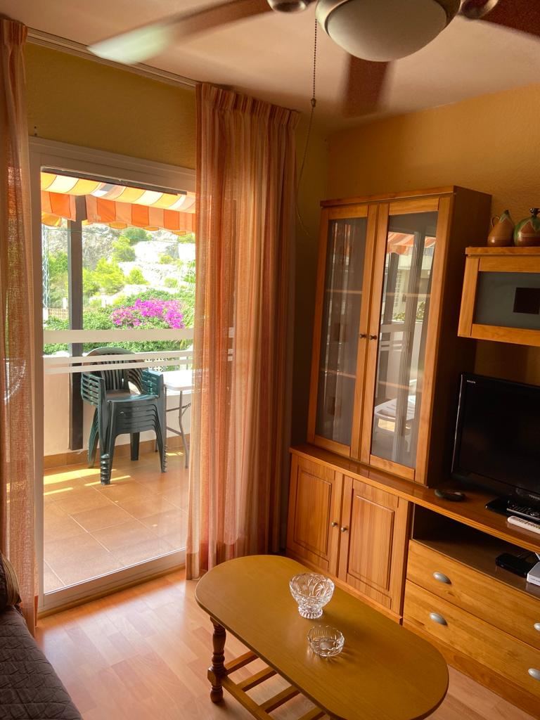 A cozy living area features a wooden cabinet displaying decor items and a television. A small table sits in front, while a ceiling fan offers airflow. Natural light filters through sheer curtains, revealing a view of a terrace with green chairs and blooming flowers beyond a glass door.