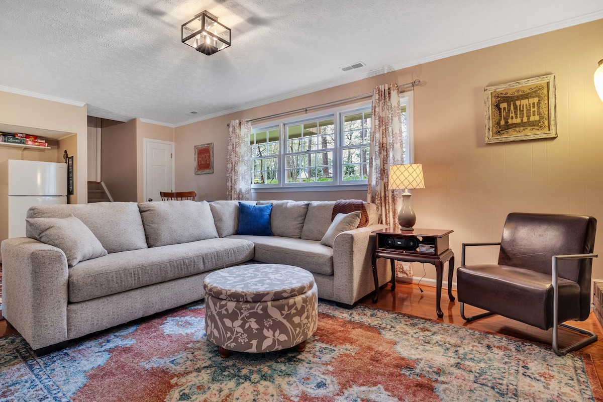 A comfortable living area is furnished with a large sectional sofa and a round ottoman, featuring a fabric pattern. A sleek, dark chair complements the space. Natural light fills the room through multiple windows. A small table with a lamp adds warmth, while a framed sign decorates the wall.