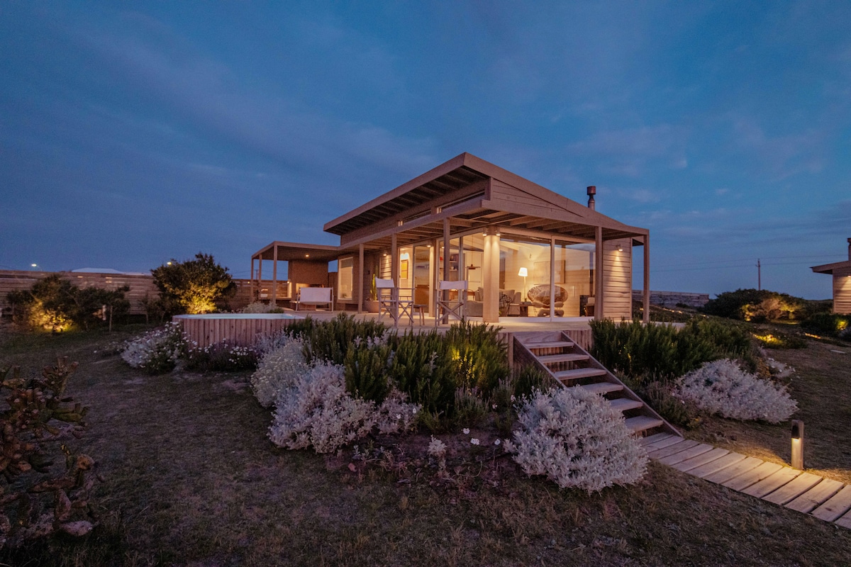 A wooden house is set against a twilight sky, featuring large glass windows that allow light to spill from within. Surrounding gardens are filled with low shrubs and flowers, while a wooden pathway leads up to the entrance, softly illuminated by ground lights.