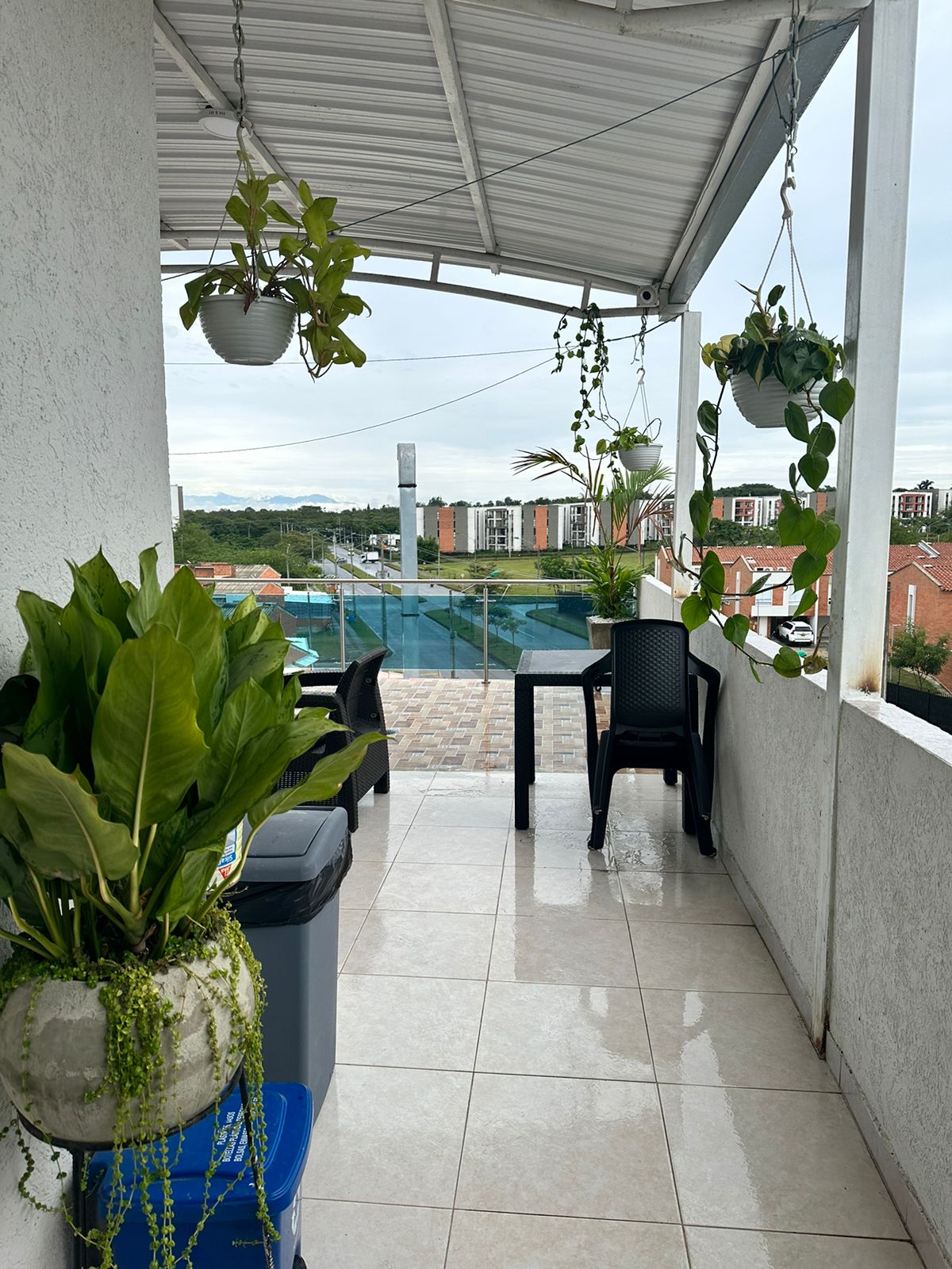 A balcony space is presented with a tiled floor, surrounded by potted plants. A black chair and table set are positioned on one side, while a trash bin is located nearby. A view of a swimming pool and neighboring buildings is visible in the background.