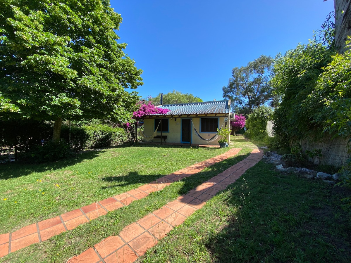 A charming house sits surrounded by lush greenery, with vibrant pink flowering plants enhancing the landscape. A winding pathway made of clay tiles leads to the entrance, framed by trees that provide shade under a clear blue sky.