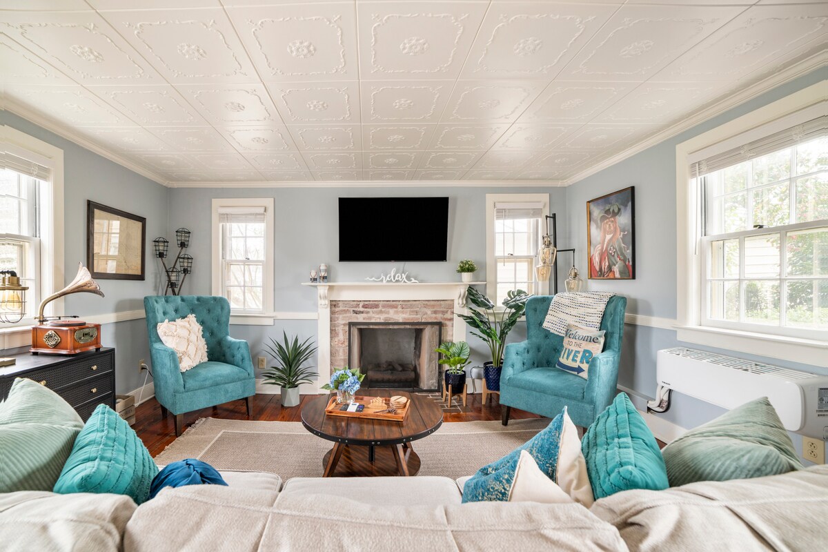 A cozy living area features two turquoise armchairs flanking a decorative fireplace. The space is brightened by natural light through multiple windows. A circular wooden table sits at the center, surrounded by green plants and decorative cushions on a light-colored rug.