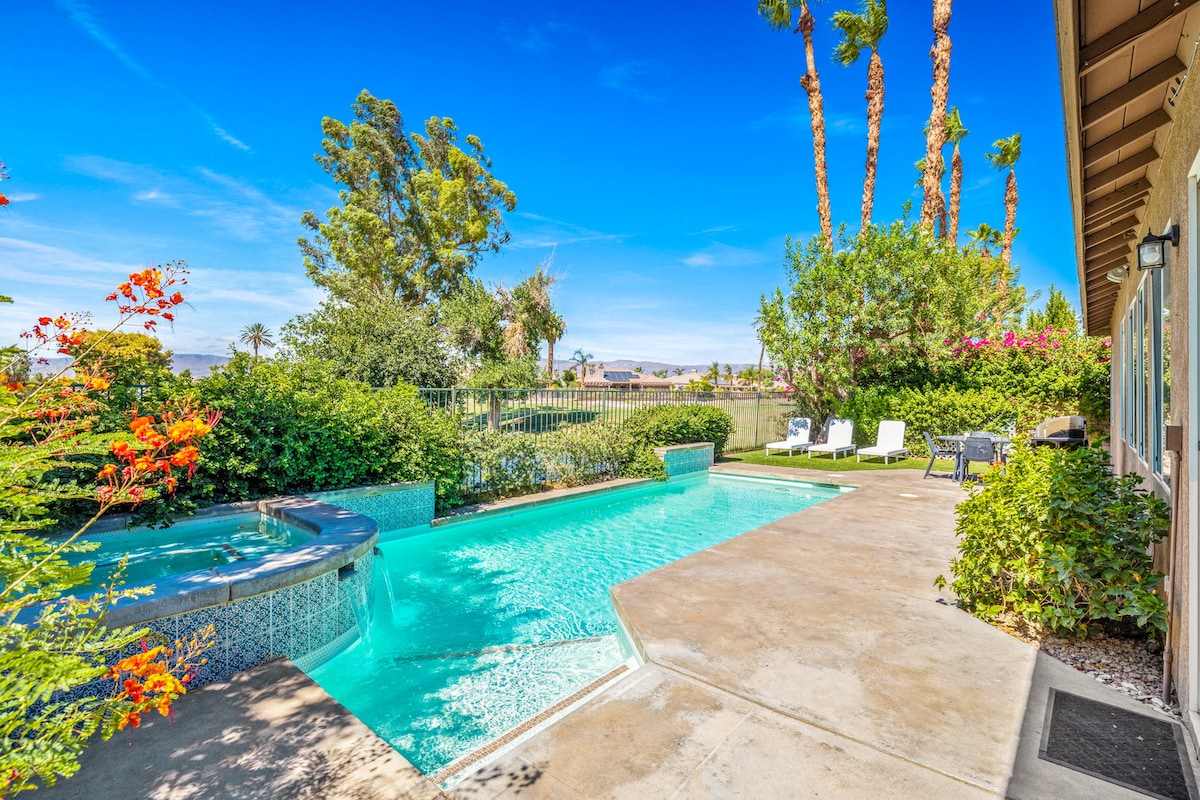 A private pool and spa are nestled within a landscaped backyard, featuring vibrant foliage and flowering plants. Loungers are positioned nearby, offering a space for relaxation, while palm trees stand tall against a bright blue sky, creating a serene outdoor atmosphere.