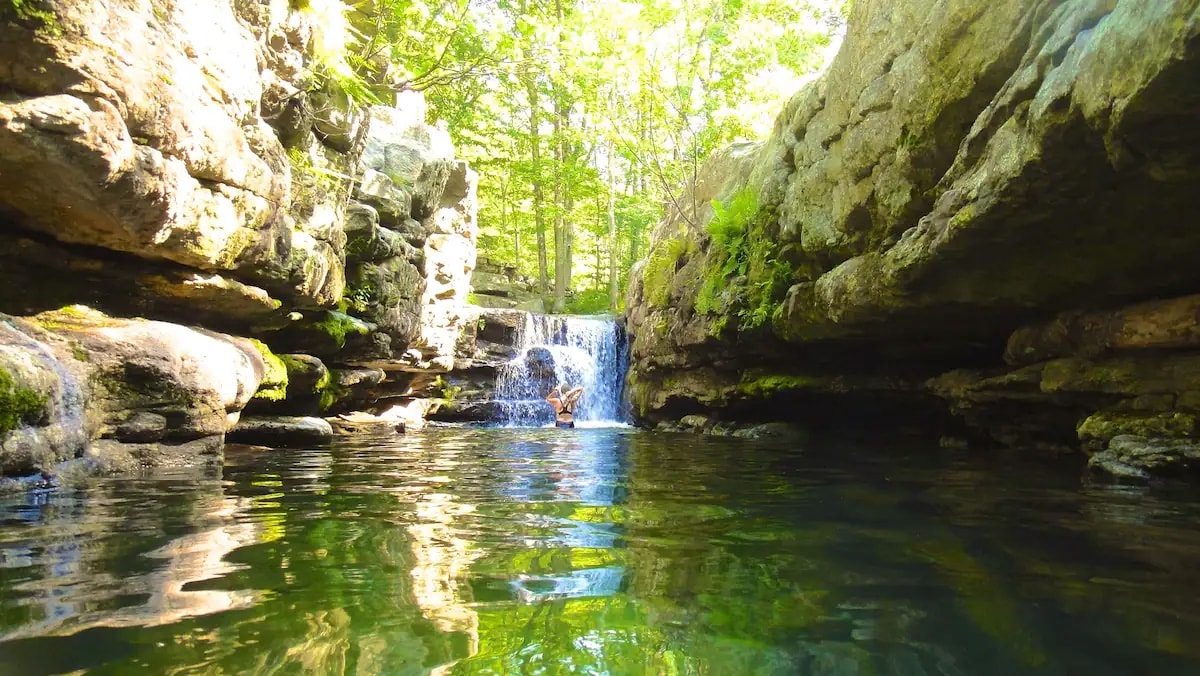 A serene natural setting is depicted, featuring smooth rocks framing a tranquil water pool. A small waterfall cascades gently into the clear water, surrounded by lush greenery. Sunlight filters through the trees, illuminating the peaceful scene.