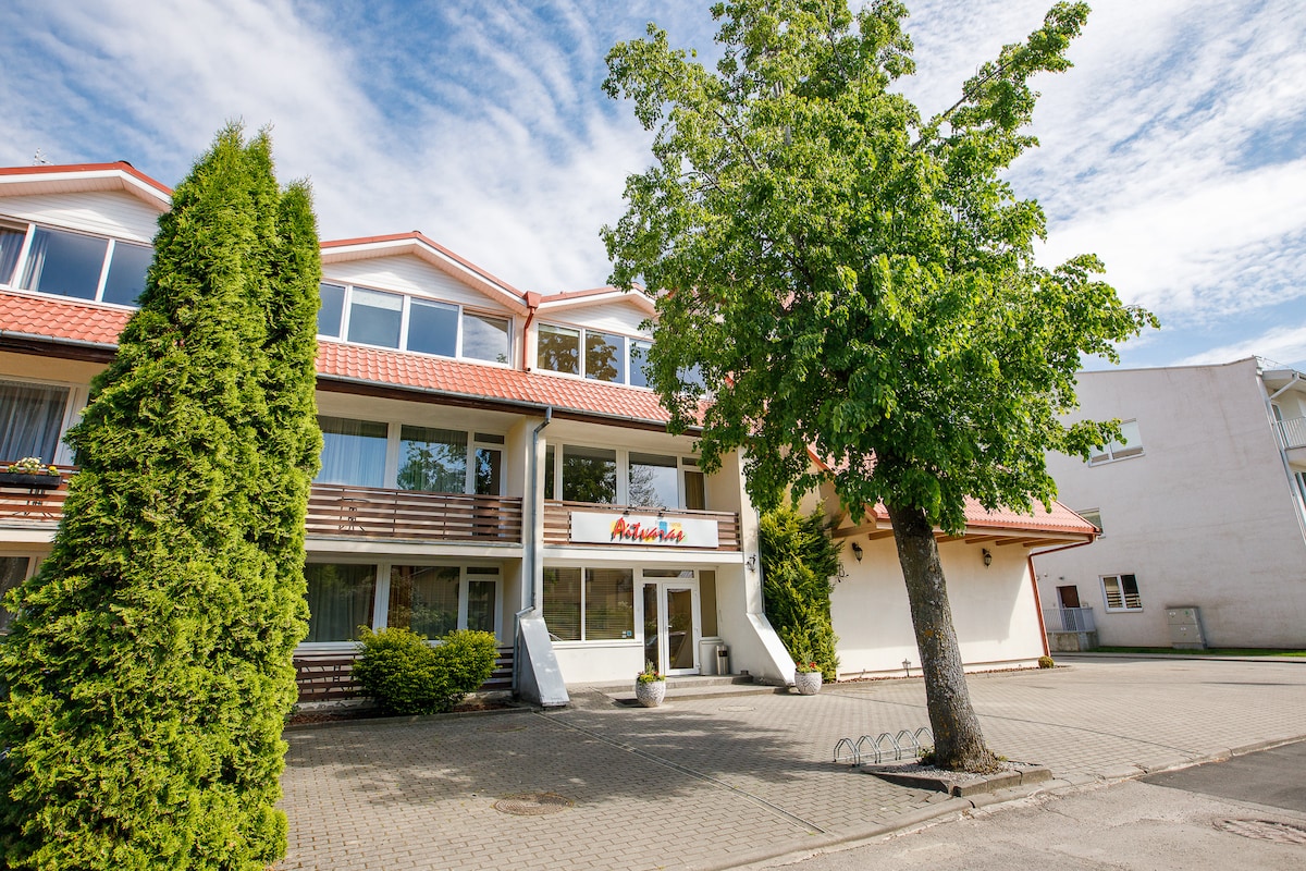 A welcoming hotel exterior features a light-colored facade with multiple large windows. A prominent tree stands in front, providing shade. The entrance is marked by a sign, and a paved area accommodates bicycles. A bright sky enhances the overall appearance.