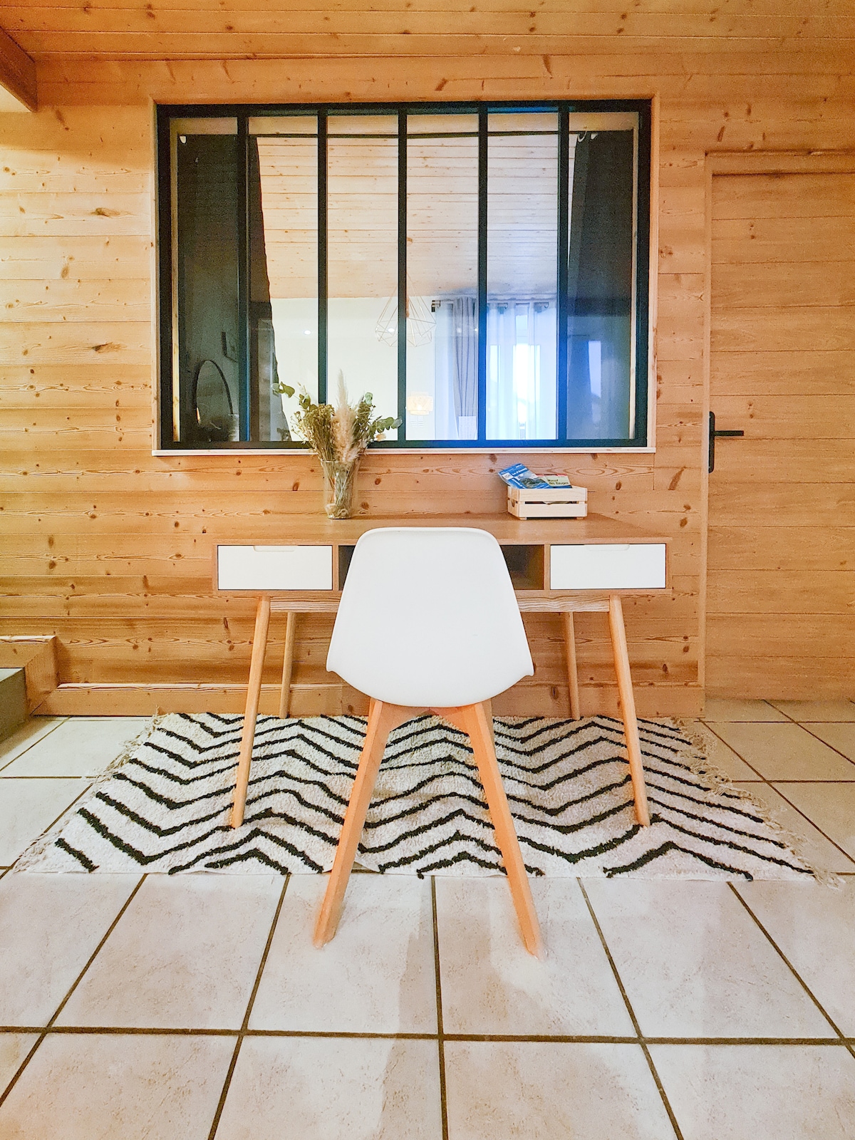 A workspace is presented with a minimalist white desk and a modern chair, set on a patterned rug. Natural wood walls provide warmth, while large windows allow ample light to illuminate the area, creating a focused atmosphere for productivity.
