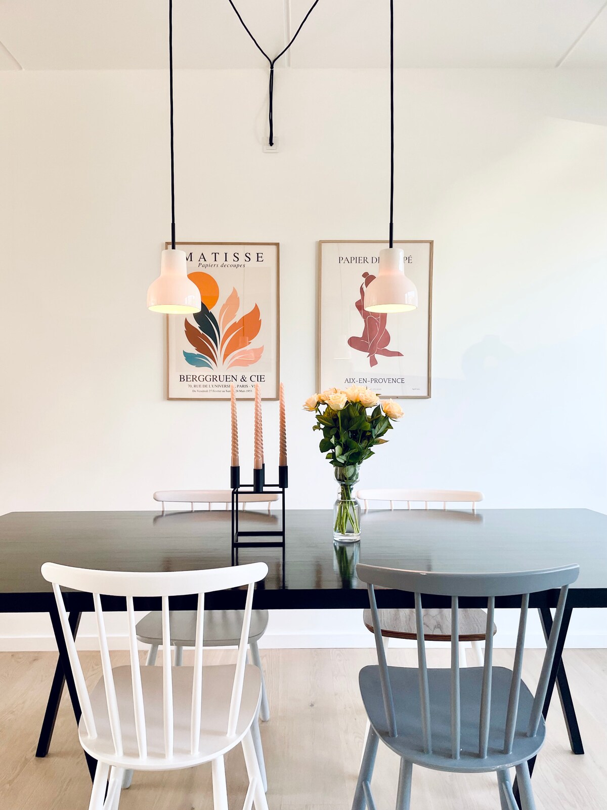 A dining area features a sleek black table surrounded by two chairs—one white and one gray. Two framed art pieces adorn the wall, complementing the space. A bouquet of fresh flowers is centered on the table, with slim candles displayed in a candle holder.
