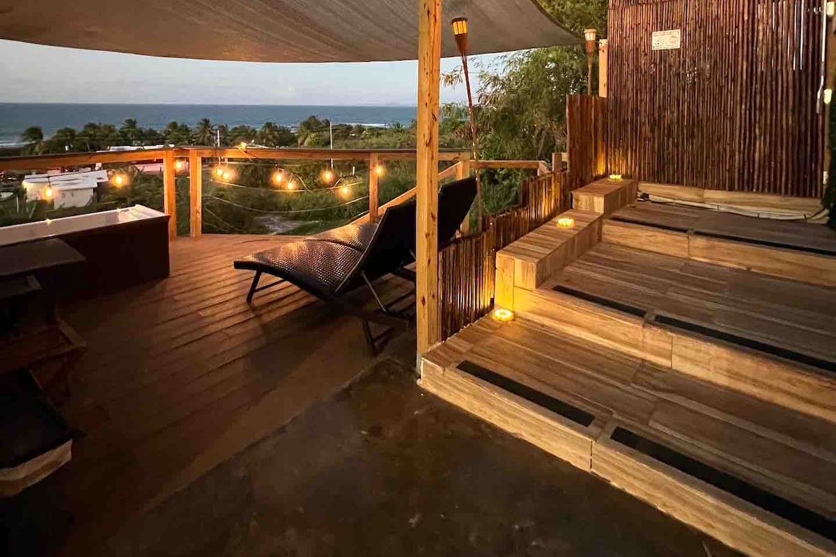 A private deck is showcased, featuring a lounge chair positioned near the edge. Soft lighting illuminates the space, highlighting the steps leading down to the jacuzzi area. The ocean view is visible beyond the greenery, creating a serene outdoor setting.