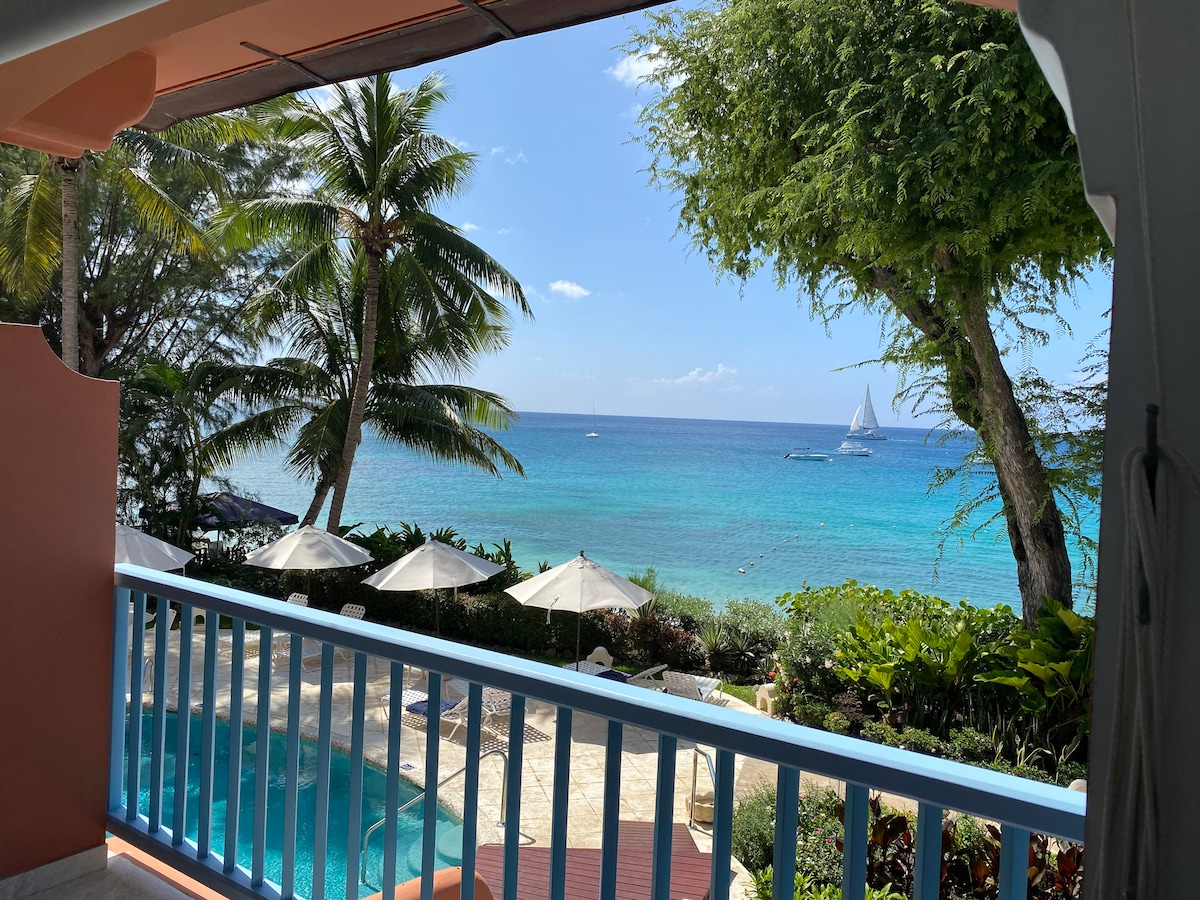A serene view is presented from a balcony, showcasing clear turquoise waters of the Caribbean Sea. Sailboats can be seen in the distance, complemented by palm trees and umbrellas by the pool below. Lush greenery frames the scene, enhancing the natural beauty.