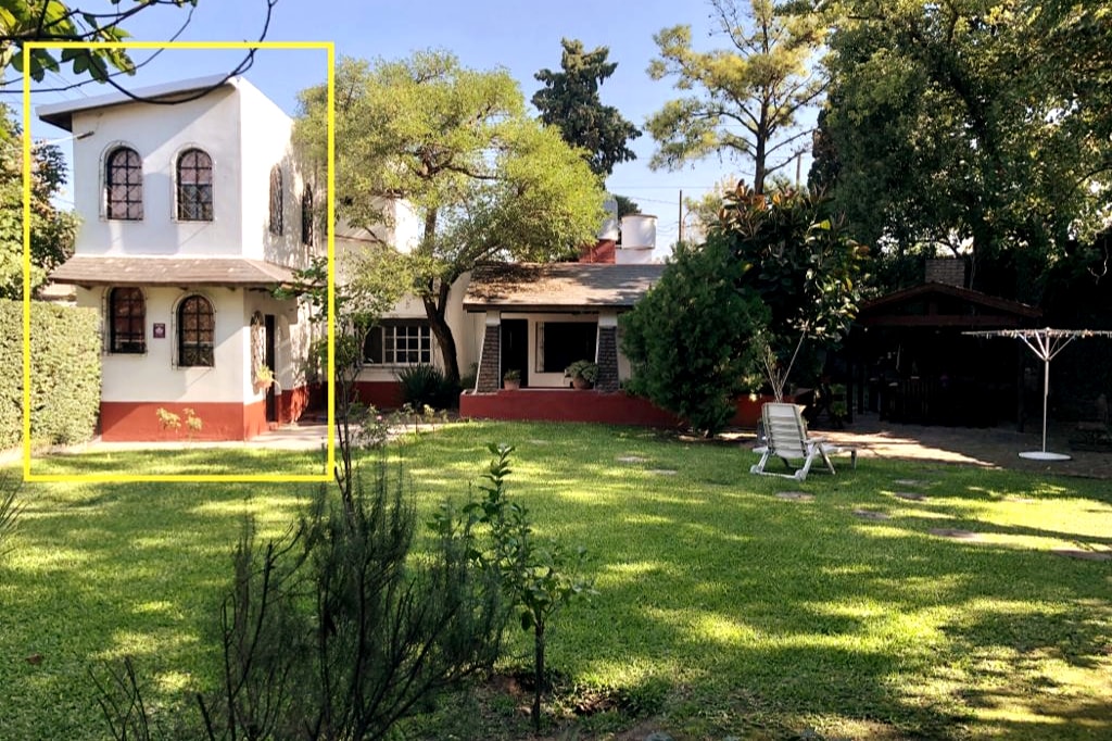 A two-story house with a white exterior and a gabled roof is visible. The front porch features a small railing, and potted plants are arranged near the entrance. The surroundings include greenery and a pathway leading to the main door.