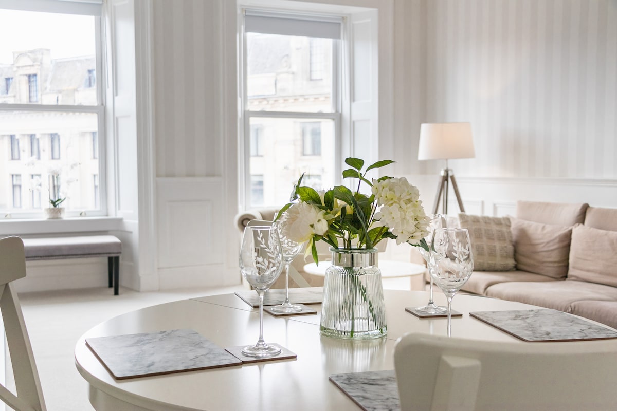 A bright dining area features a round table set with marble placemats and wine glasses, complemented by a vase of fresh flowers. Light filters through large windows, illuminating the surrounding soft furnishings and inviting atmosphere.