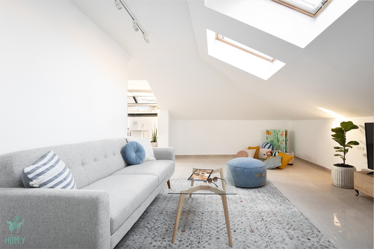 A spacious living area features a light grey sofa adorned with a blue pillow, complemented by a round blue ottoman. Natural light floods the space through skylights, illuminating a patterned area rug and a small potted plant in the corner. Art pieces are subtly displayed on the wall.