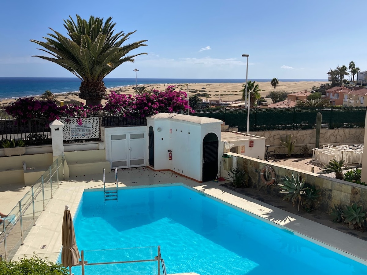 A well-maintained heated pool is surrounded by tropical plants and palm trees, providing a relaxing atmosphere. The ocean and sandy dunes are visible in the background, with a clear sky overhead. Access to the pool area is unobstructed, enhancing the coastal experience.