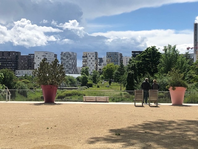 A scenic view of a tranquil park is presented, featuring greenery and modern buildings in the background. Two individuals are seen sitting on benches, while large potted plants add structure to the landscape. The sky is partly cloudy, enhancing the natural ambiance.