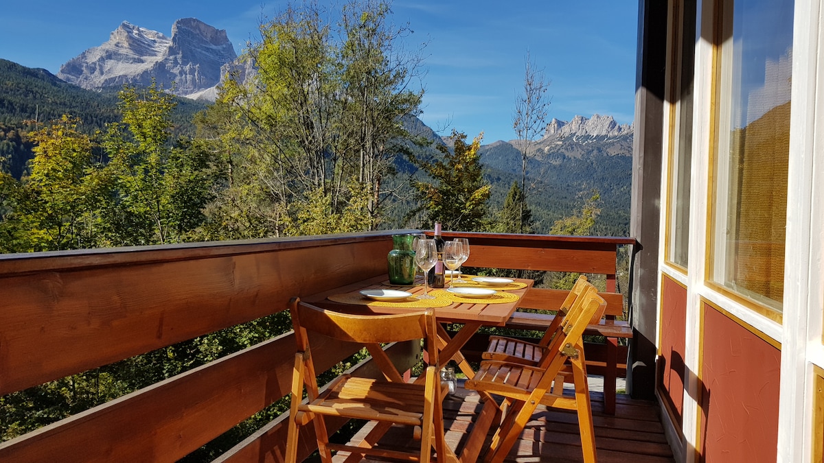 An outdoor balcony featuring a wooden table set for two, complete with glasses and plates. The surrounding trees provide a natural backdrop, while the stunning mountains rise majestically in the distance under a clear blue sky.