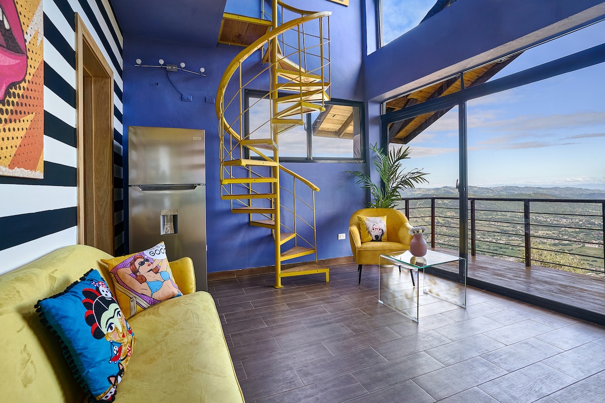 A vibrant loft space features a spiral staircase leading to upper levels. A mustard-yellow armchair and a colorful sofa are positioned next to a glass coffee table. Floor-to-ceiling windows provide scenic views, while a minimalist kitchen area is visible in the background.